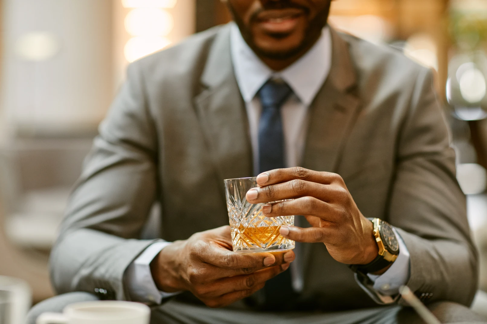 A finely appointed Black man weating a gray suit, blue tie and expensive watch holding a crystal glass with a whiskey beverage while seated in a clubroom.