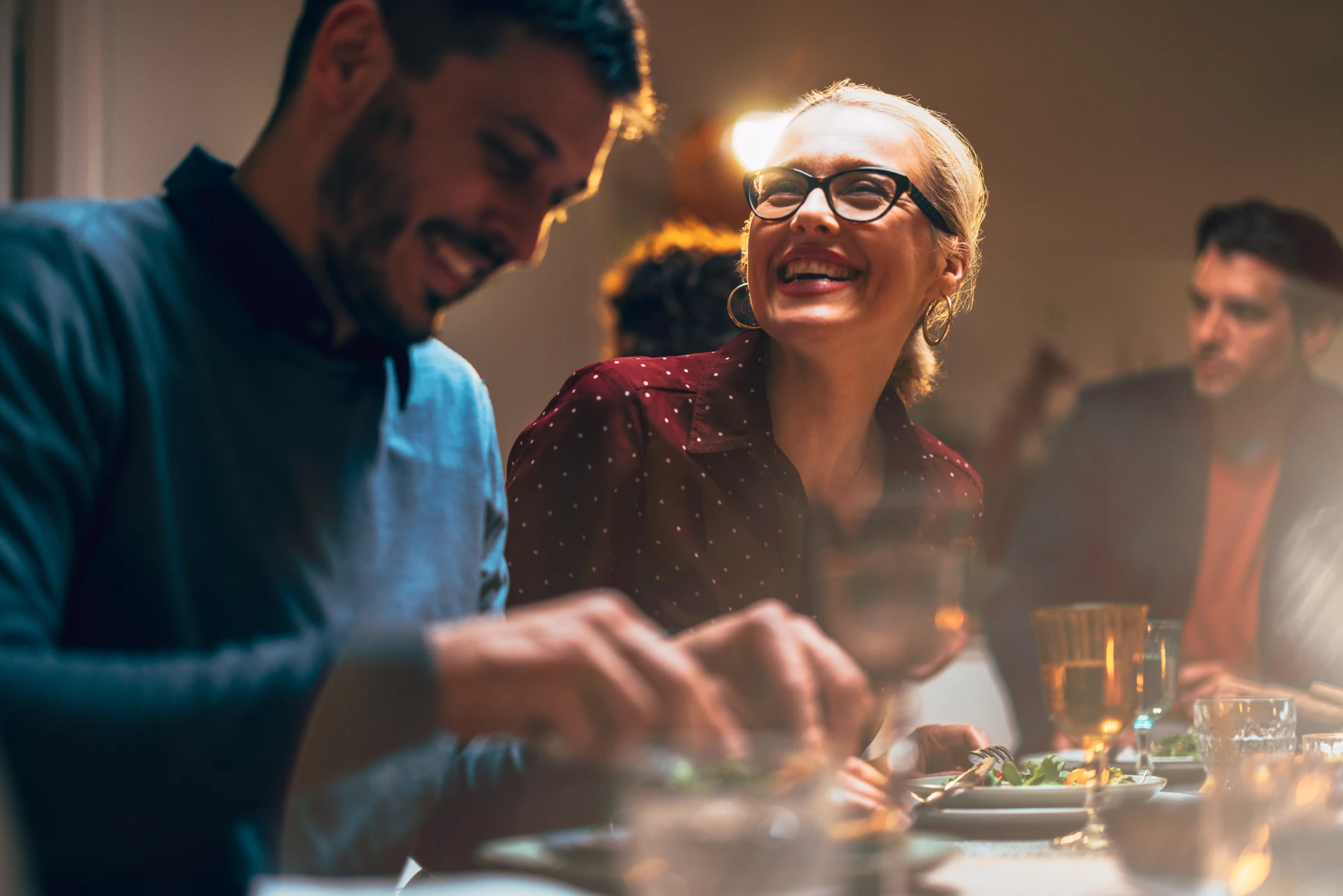 A young, bearded man and a laughing woman wearing glasses enjoying salads for dinner in a dimly lit and bustling downtown restaurant.